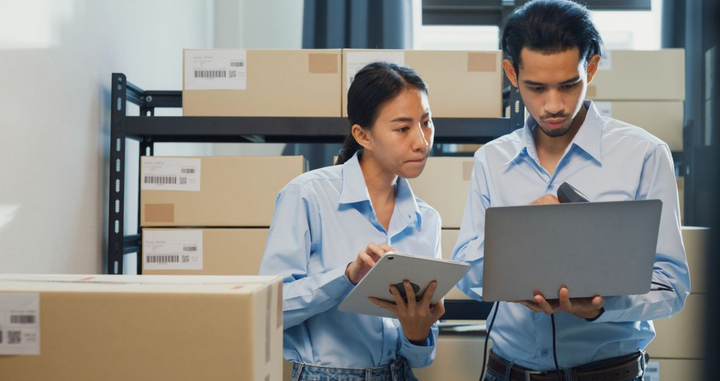 Empleados revisando información en un portátil y una tablet en un almacén, gestionando inventario en tiempo real.