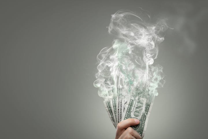 Una mano sosteniendo varios billetes de dólar estadounidense disolviéndose en el aire, asemejándose al humo.
